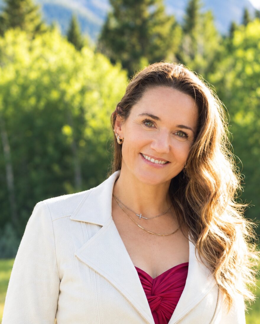 Julia McElgunn smiling, in a white jacket with aspen trees behind her